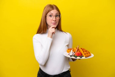 Sarı arka planda izole edilmiş waffle tutan genç kızıl saçlı kadının şüpheleri ve düşünceleri var.
