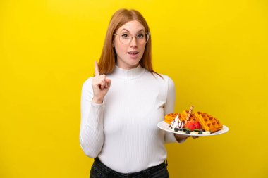Sarı arka planda izole edilmiş waffle 'ları tutan genç kızıl saçlı kadın bir fikrin işaret ettiğini düşünüyor.