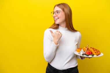 Sarı arka planda izole edilmiş waffle tutan genç kızıl saçlı kadın bir ürünü sunmak için yan tarafı işaret ediyor.