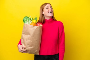 Sarı arka planda izole edilmiş alışveriş torbası tutan genç kızıl saçlı kadın gülüyor.