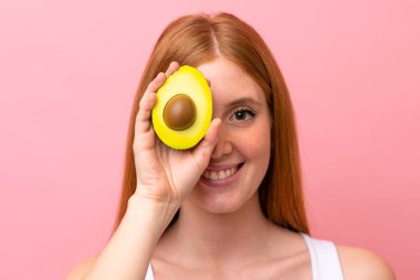 Genç kızıl saçlı kadın pembe arka planda izole edilmiş, elinde avakado tutarken gülümsüyor. Portreyi kapat