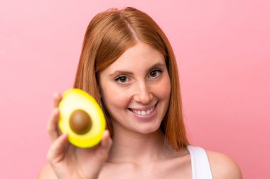 Genç kızıl saçlı kadın pembe arka planda izole edilmiş, elinde avakado tutarken gülümsüyor. Portreyi kapat