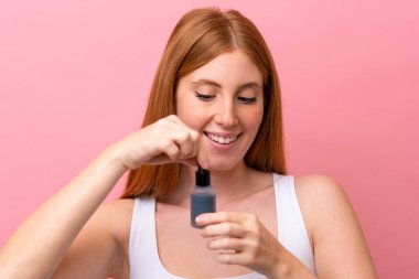 Genç kızıl saçlı kadın pembe arka planda izole edilmiş, elinde bir serum tutarken gülümsüyor. Portreyi kapat