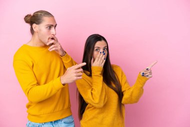 Pembe arka planda izole edilmiş genç beyaz çift şaşırmış bir yüzle parmaklarını yana gösteriyor.