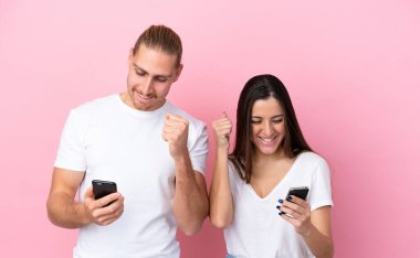 Pembe arka planda izole edilmiş genç beyaz bir çift cep telefonu kullanıyor ve zafer jesti yapıyor.