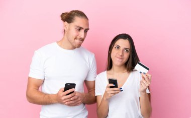 Kafkasyalı genç çift pembe arka planda izole edilmiş. Cep telefonuyla kredi kartıyla alışveriş yapıyorlar.