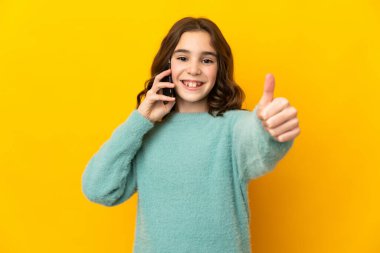Sarı arka planda izole edilmiş küçük beyaz kız başparmaklarını kaldırırken cep telefonuyla sohbet ediyor.