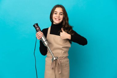 El blenderi kullanan küçük bir kız mavi arka planda izole edilmiş bir şekilde baş parmak hareketi yapıyor.