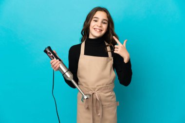 El blenderi kullanan küçük bir kız mavi arka planda izole edilmiş bir şekilde baş parmak hareketi yapıyor.