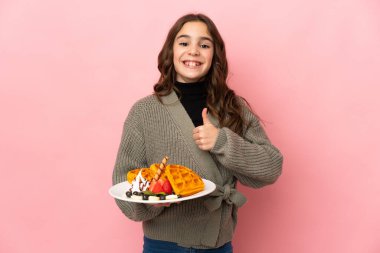 Pembe arka planda izole edilmiş waffle 'lar tutan küçük bir kız baş parmak işareti yapıyor.