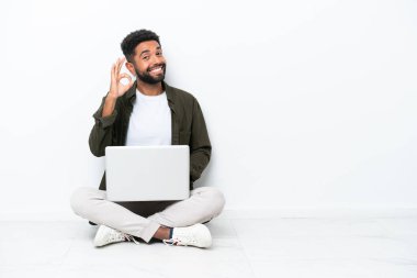 Yerde dizüstü bilgisayarı olan genç Brezilyalı bir adam izole bir şekilde beyaz üzerinde oturuyor. Parmaklarıyla tamam işareti gösteriyor.