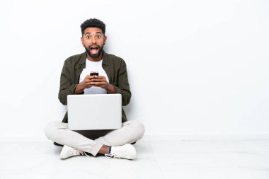 Yerde dizüstü bilgisayarlı genç Brezilyalı bir adam oturuyor. Beyazlara izole edilmiş, şaşırmış ve mesaj yolluyor.