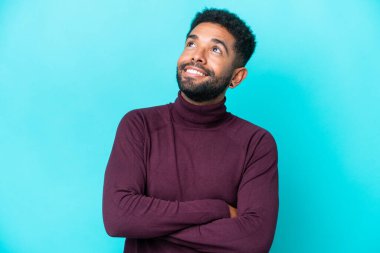 Genç Brezilyalı adam mavi arka planda izole edilmiş gülümserken yukarı bakıyor