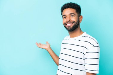 Mavi arka planda izole edilmiş genç Brezilyalı. Davet etmek için ellerini yana uzatıyor.