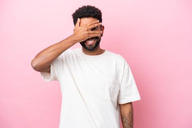 Pembe arka planda izole edilmiş genç Brezilyalı adam gözlerini elleriyle kapatıp gülümsüyor.