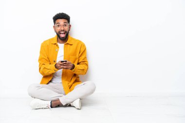 Yerde oturan Brezilyalı genç adam beyaz arka planda izole edilmiş, şaşırmış ve bir mesaj gönderiyor.