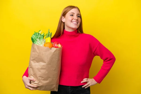 若いです赤毛女性保持しています食料品のショッピングバッグ孤立した上に黄色の背景ポージングとともに腕でヒップと笑顔 — ストック写真