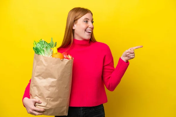 Jonge Roodharige Vrouw Met Een Boodschappentas Geïsoleerd Gele Achtergrond Wijzende — Stockfoto