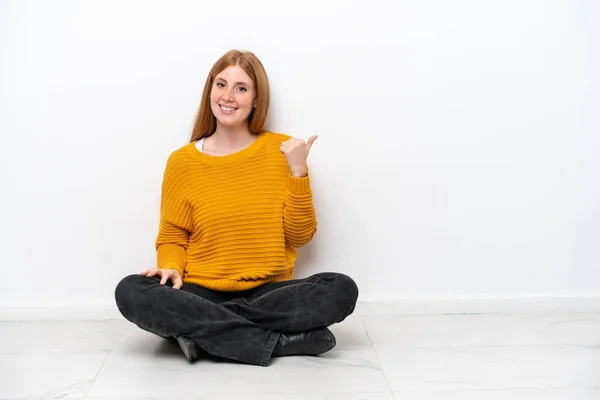 一个红头发的年轻女人坐在地板上 被白色的背景隔离着 指着旁边展示着一种产品 — 图库照片