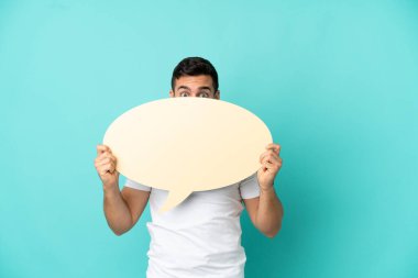 Mavi arka planda izole edilmiş yakışıklı beyaz adam arkasında saklanan boş bir konuşma balonu tutuyor.