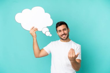 Genç, beyaz, mavi arka planda izole edilmiş bir adam elinde bir konuşma balonu tutuyor ve gelecek vadeden bir jest yapıyor.