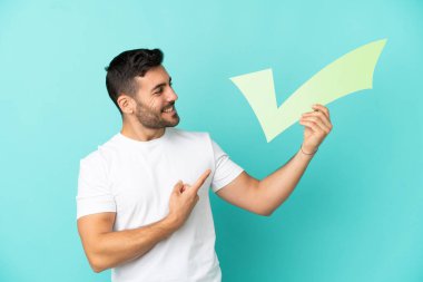Mavi arka planda izole edilmiş yakışıklı beyaz bir adam elinde bir çek simgesi tutuyor ve ona doğrultuyor.