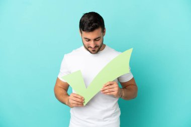 Mavi arka planda izole edilmiş yakışıklı beyaz adam elinde bir çek simgesi tutuyor ve ona bakıyor.