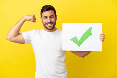 Sarı arka planda izole edilmiş yakışıklı beyaz adam elinde yeşil işaret simgesi olan bir levha tutuyor ve güçlü bir hareket yapıyor.