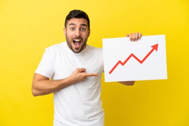 Sarı arka planda izole edilmiş yakışıklı beyaz bir adam elinde büyüyen bir istatistik okuyla şaşırmış bir ifadeyle bir işaret tutuyor.