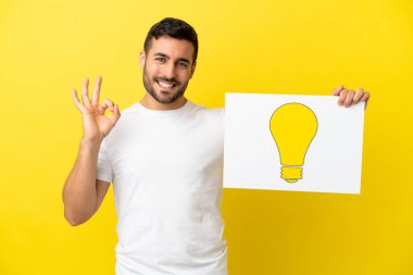 Sarı arka planda izole edilmiş yakışıklı beyaz adam elinde üzerinde ok işareti olan ampul simgesi olan bir levha tutuyor.