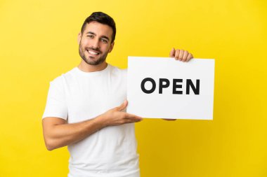 Sarı arka planda izole edilmiş yakışıklı beyaz bir adam elinde mutlu bir ifadeyle barış sembolü taşıyan bir levha tutuyor.