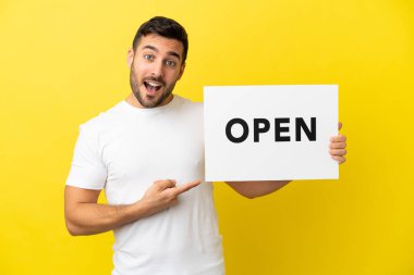 Sarı arka planda izole edilmiş yakışıklı beyaz bir adam. Elinde açık yazılı bir levha var ve onu gösteriyor.