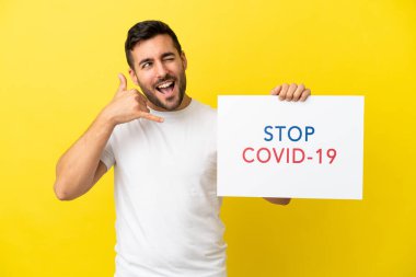Sarı arka planda izole edilmiş yakışıklı beyaz bir adam elinde yazılı bir levhayla Covid 19 'u durdur ve telefon hareketi yap.