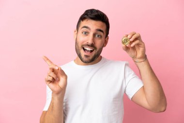 Elinde pembe arka planda izole edilmiş bir Bitcoin tutan genç adam, parmağıyla kenarı işaret ediyor.