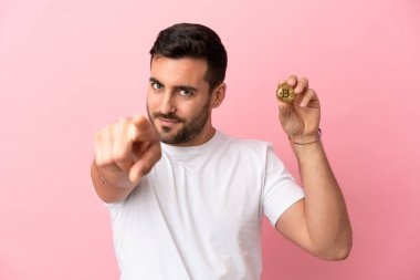 Elinde pembe arka planda izole edilmiş bir Bitcoin tutan genç adam mutlu bir ifadeyle ön tarafı işaret ediyor.