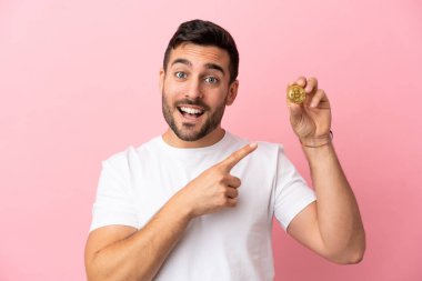 Elinde pembe arka planda izole edilmiş bir Bitcoin tutan genç adam bir ürünü sunmak için kenarı işaret ediyor.