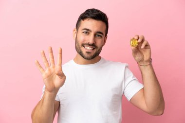Elinde pembe arka planda izole edilmiş Bitcoin tutan genç adam mutlu ve parmaklarıyla dörde kadar sayıyor.