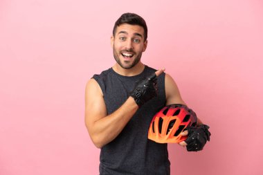Pembe arka planda izole edilmiş genç bisikletçi bir ürünü sunmak için kenarı işaret ediyor.
