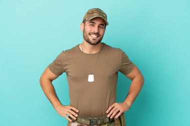 Mavi arka planda izole edilmiş, kolları kalçasında ve gülümseyerek poz veren asker.