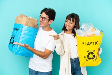 Mavi arka planda izole edilmiş plastik ve kâğıt dolu bir poşet taşıyan genç çift geriye dönüp bir ürün sunuyor.