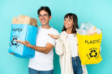 Mavi arka planda izole edilmiş plastik ve kâğıt dolu bir çanta taşıyan genç çift, davete gelmek için ellerini yana uzatıyor.