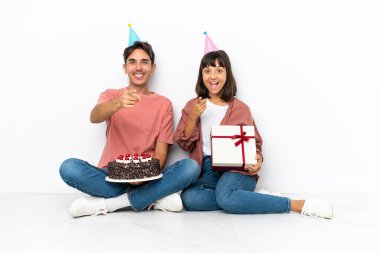 Genç melez çift, beyaz arka planda izole bir şekilde yerde oturmuş doğum gününü kutluyorlar.