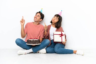 Genç melez çift, beyaz arka planda izole bir şekilde yerde oturmuş doğum gününü kutluyor. Harika bir fikre işaret ediyor ve yukarı bakıyor.