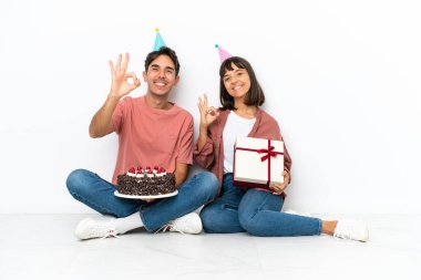 Genç melez çift, beyaz arka planda izole bir şekilde yerde oturmuş doğum gününü kutluyorlar.
