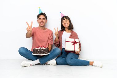 Genç melez çift, beyaz arka planda izole bir şekilde yerde oturmuş doğum günü kutluyor gülümsüyor ve zafer işaretlerini iki eliyle gösteriyor.