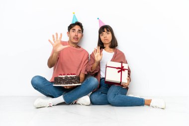 Genç melez çift, beyaz arka planda izole bir şekilde yerde oturmuş doğum gününü kutluyor. Biraz gergin ve korkmuş bir şekilde ellerini öne uzatıyor.