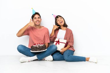 Genç melez bir çift, beyaz arka planda izole bir şekilde yerde oturmuş doğum gününü kutluyorlar ve telefon ediyorlar. Bana geri dönüş işareti ver.