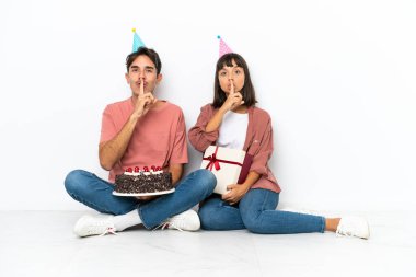 Genç melez çift, beyaz arka planda izole bir şekilde yerde oturmuş doğum gününü kutluyorlar.