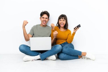 Dizüstü bilgisayarı ve cep telefonu olan genç bir çift yerde oturmuş zaferi kutluyor.