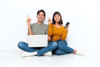 Dizüstü bilgisayarı ve cep telefonu olan genç bir çift yerde oturuyor ve en iyinin göstergesi olarak parmağını kaldırıyor.